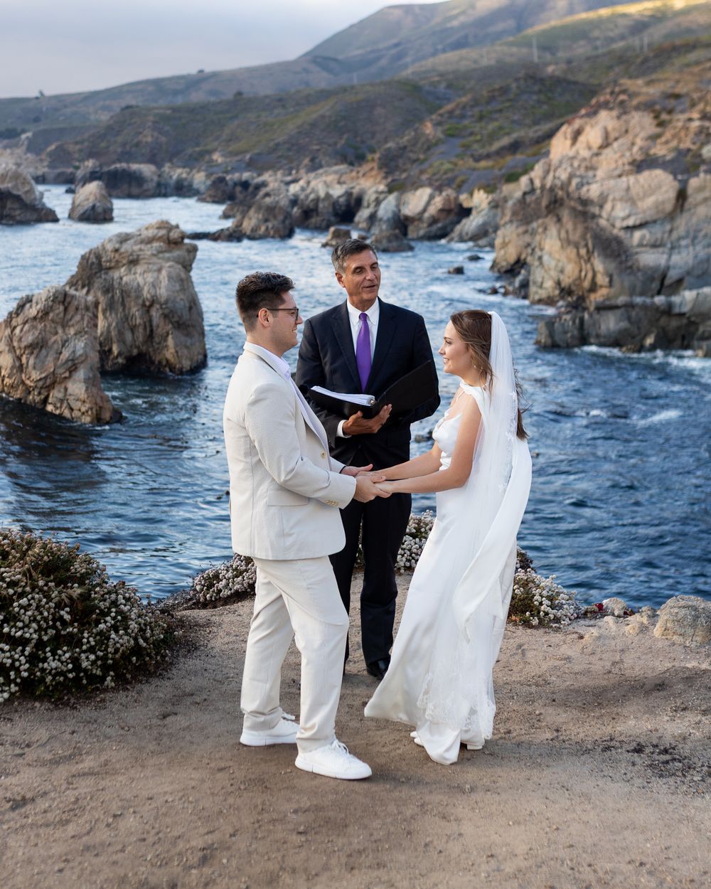A wedding ceremony takes place on a scenic coastal cliff overlooking rocky shores and blue ocean waters.