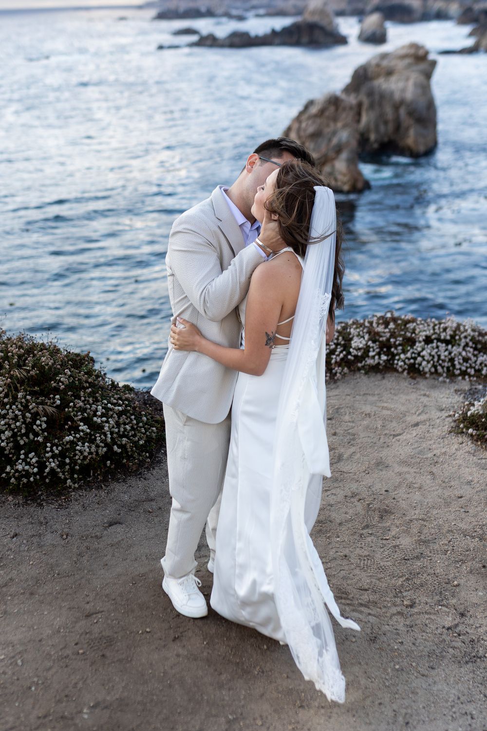Bride and groom share a romantic kiss by the ocean at sunset, bride wearing white gown and veil.
