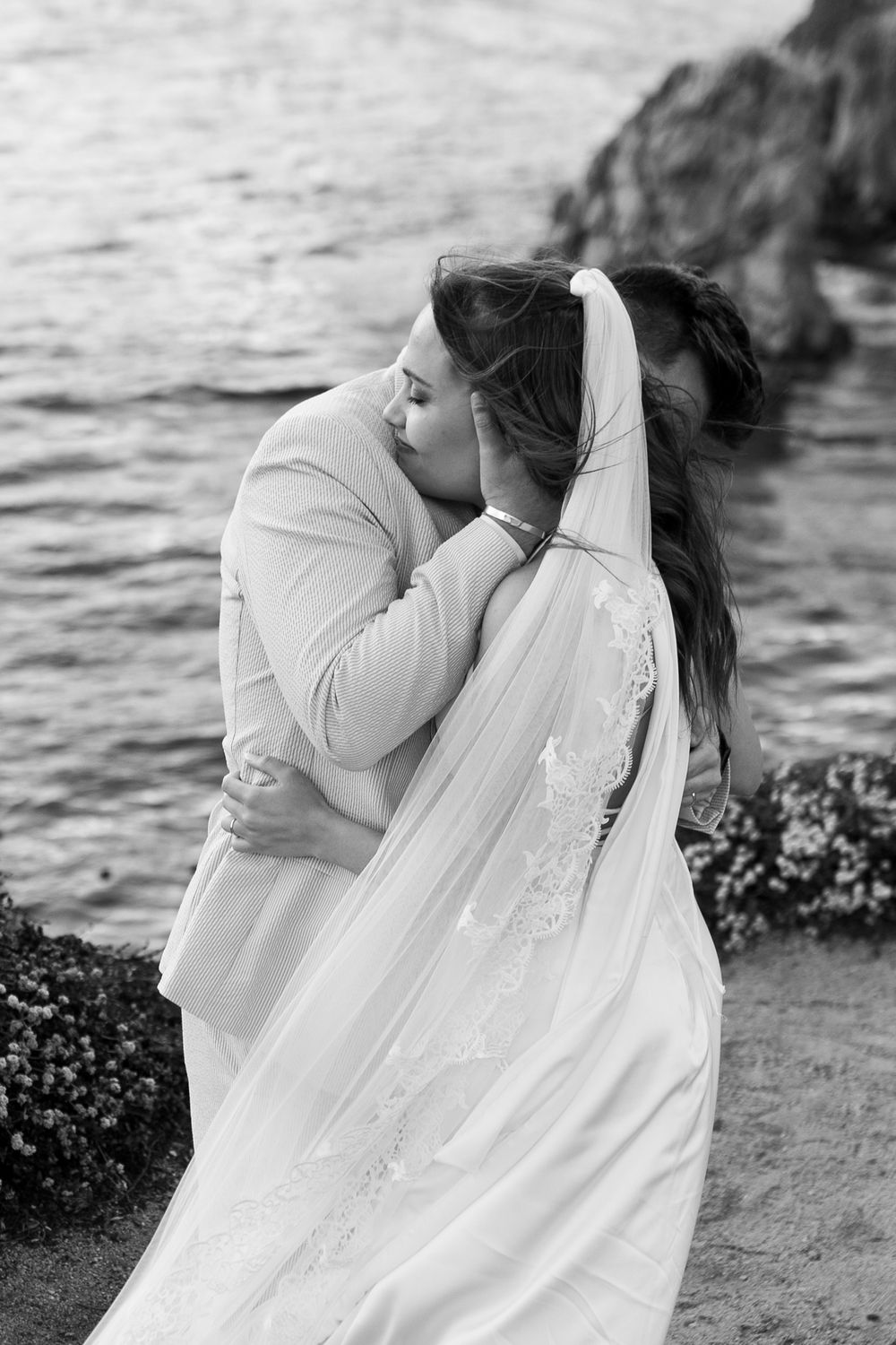 Black and white photo of a couple sharing a romantic kiss by the ocean, bride in a flowing veil and gown.