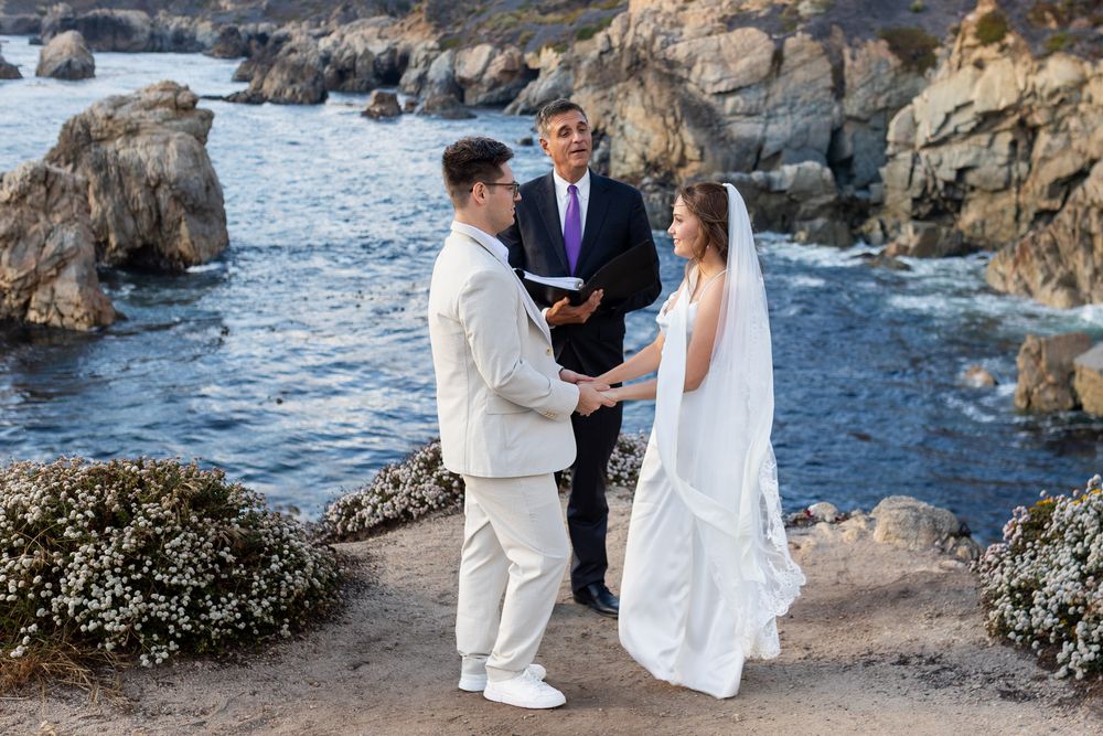 A couple shares an intimate moment during their beach wedding ceremony along a rocky coastal shoreline at sunset.