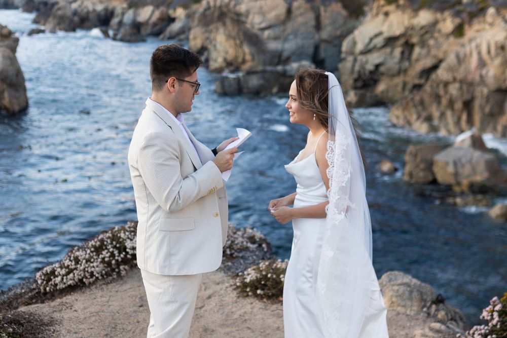 Couple exchanging vows at a cliffside ocean wedding, groom in white suit reading from paper to bride in wedding dress and veil.