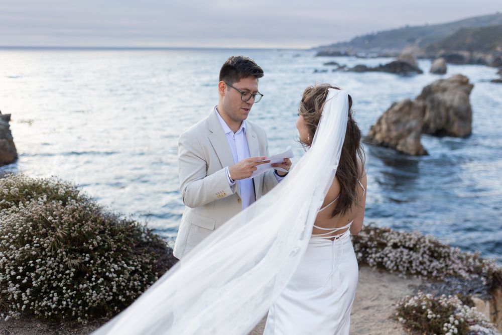 Couple exchanging vows during a romantic oceanside wedding ceremony with rocky cliffs and ocean waves in the background.