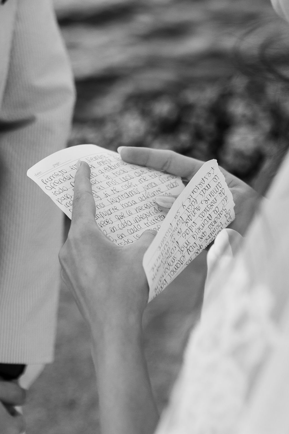 Black and white photo of hands holding handwritten wedding vows on paper during an outdoor ceremony.