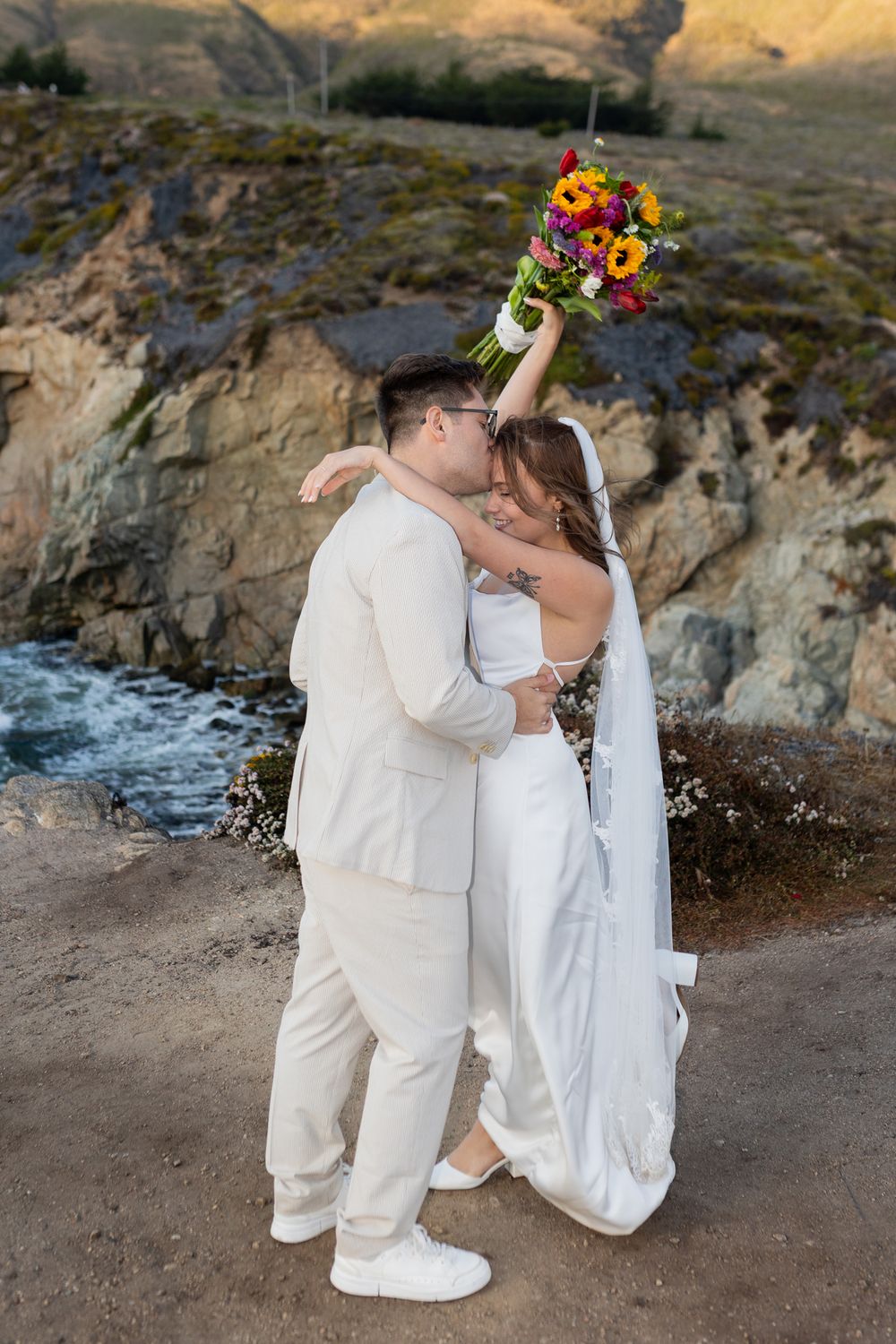 Newlywed couple kissing on rocky coastal cliffside, bride in white gown holding colorful bouquet aloft, groom in cream suit.