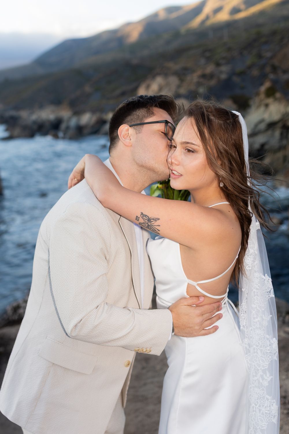 Couple in wedding attire sharing a romantic kiss by the ocean with scenic coastal mountains in the background.
