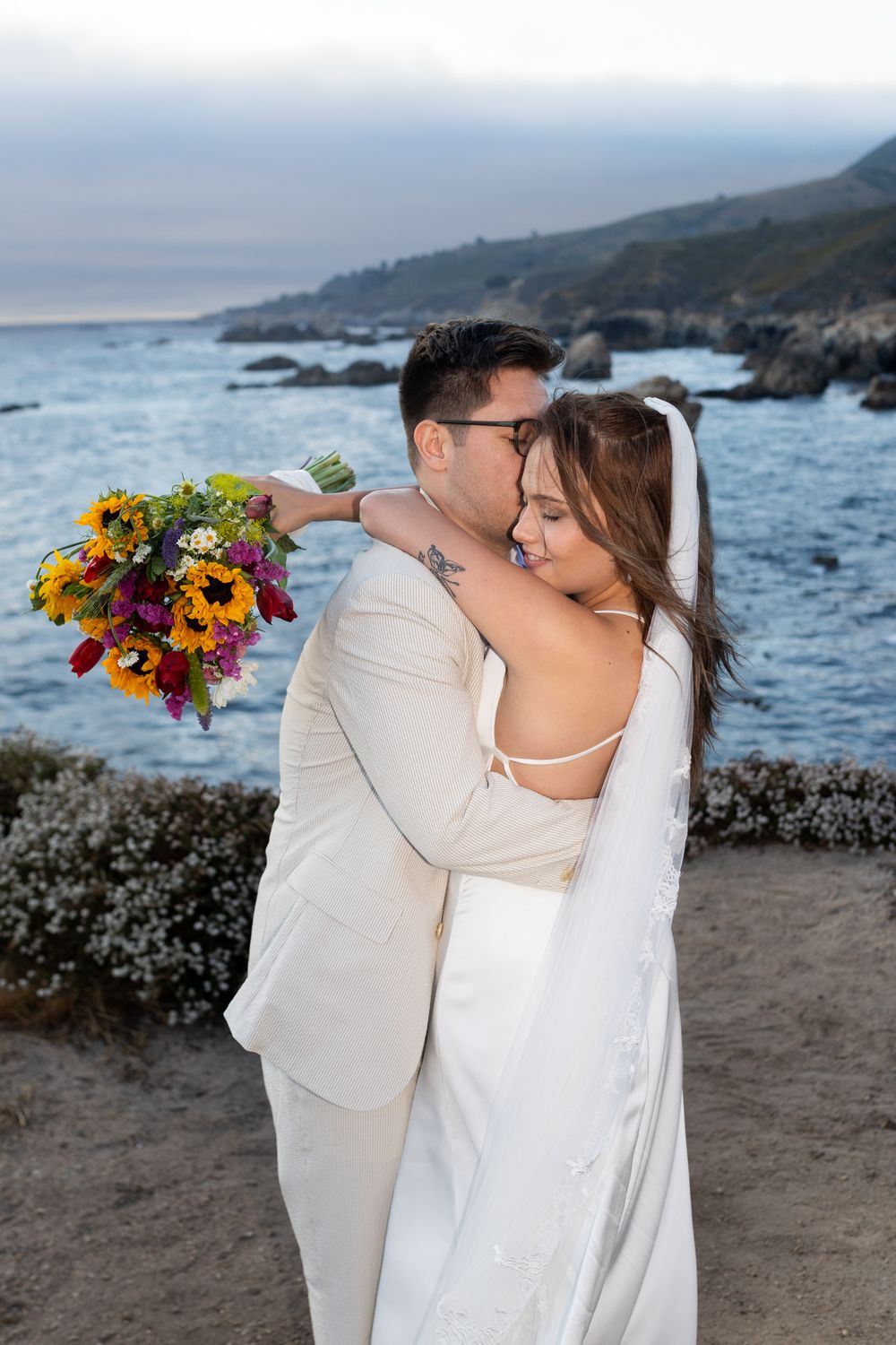 A couple in wedding attire embraces on a rocky beach with dramatic coastal cliffs and ocean views at sunset.