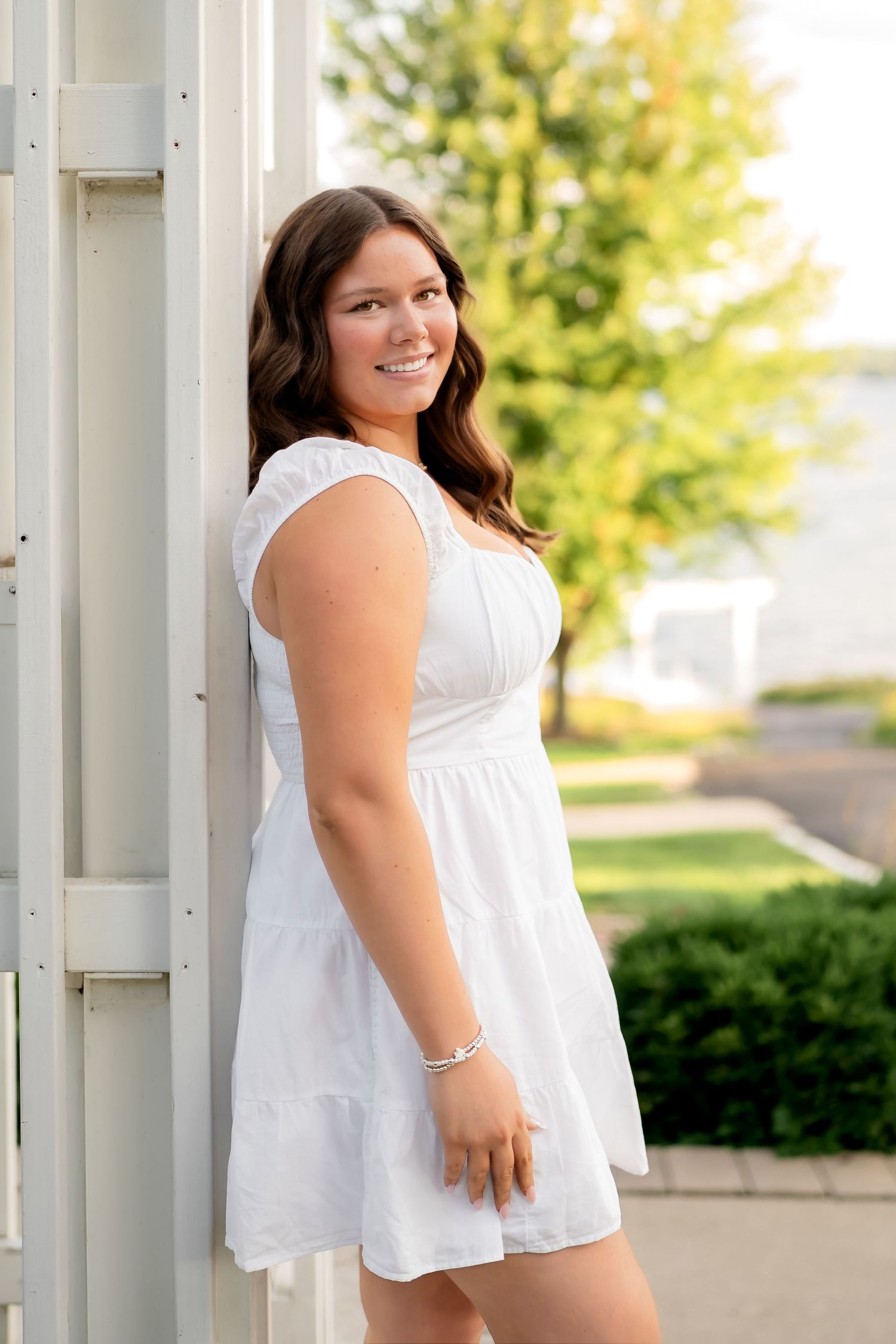 A person in a white summer dress poses outdoors against a white column with greenery in the background.