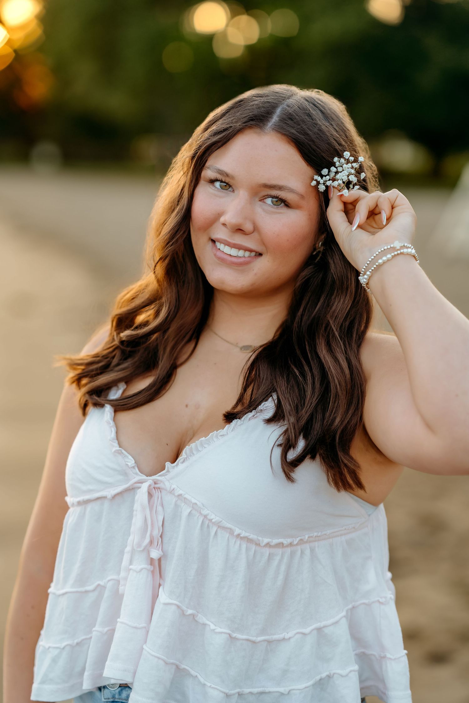 A young woman in a white tiered dress adjusts flowers in her hair during golden hour sunset lighting.