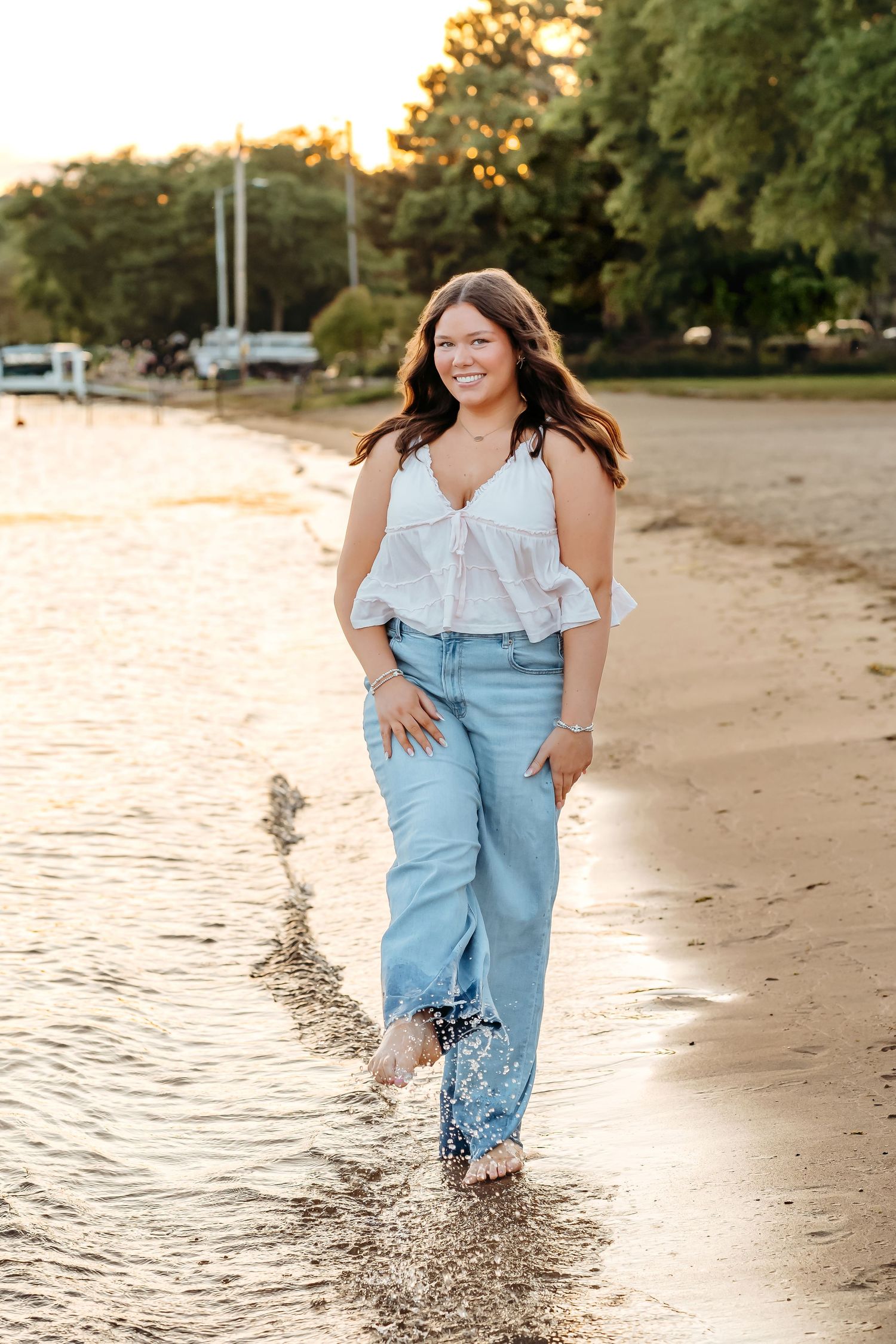 Person wearing denim overalls splashes joyfully through shallow water at the beach during golden hour.