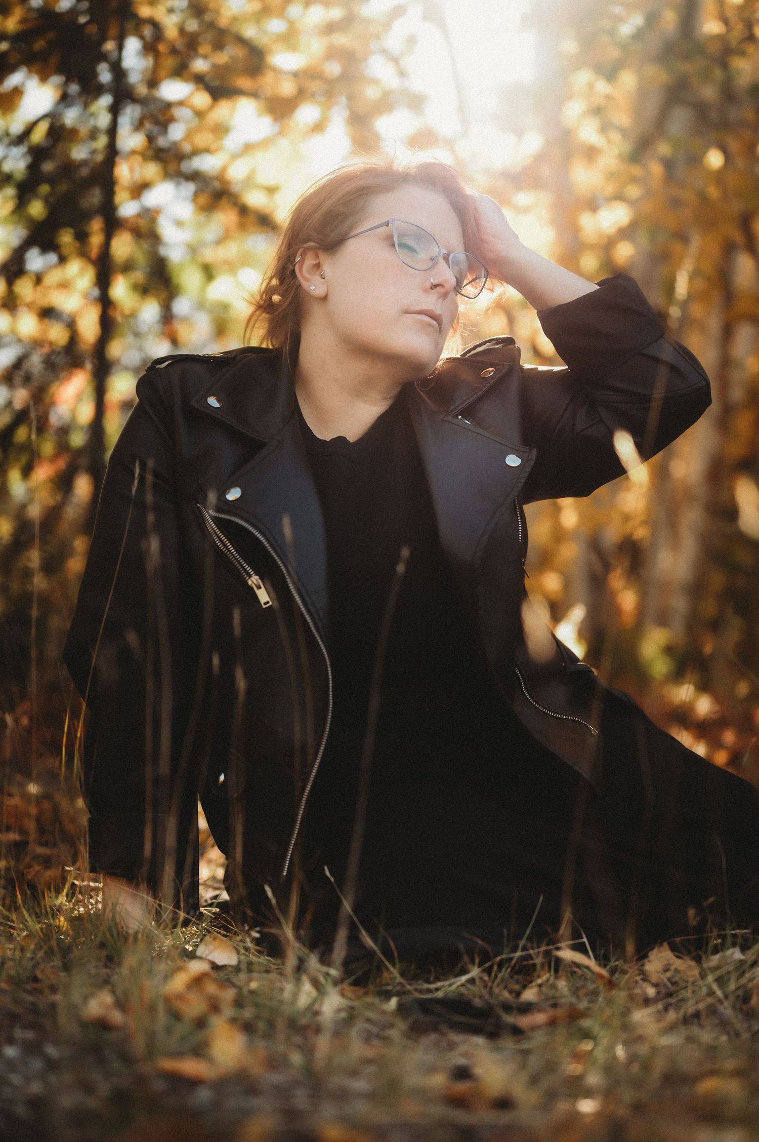 Person in black leather jacket lying on autumn ground with golden sunlight filtering through trees in background.