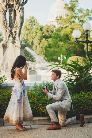 A surprise proposal taking place in front of the US Capitol with ornate fountains and lamp posts.