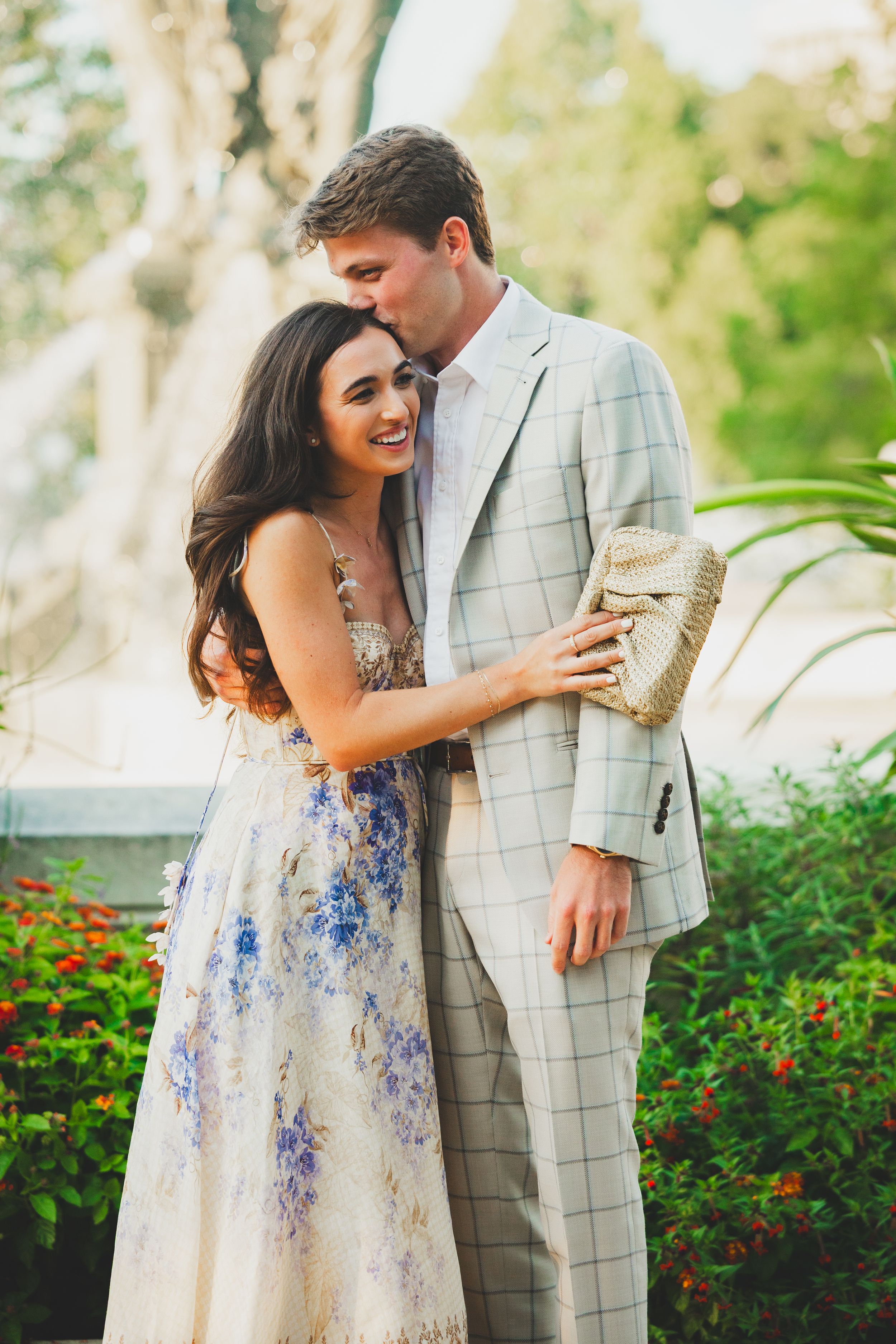 A couple in formal attire share a romantic moment in a garden setting with blooming red flowers and greenery.