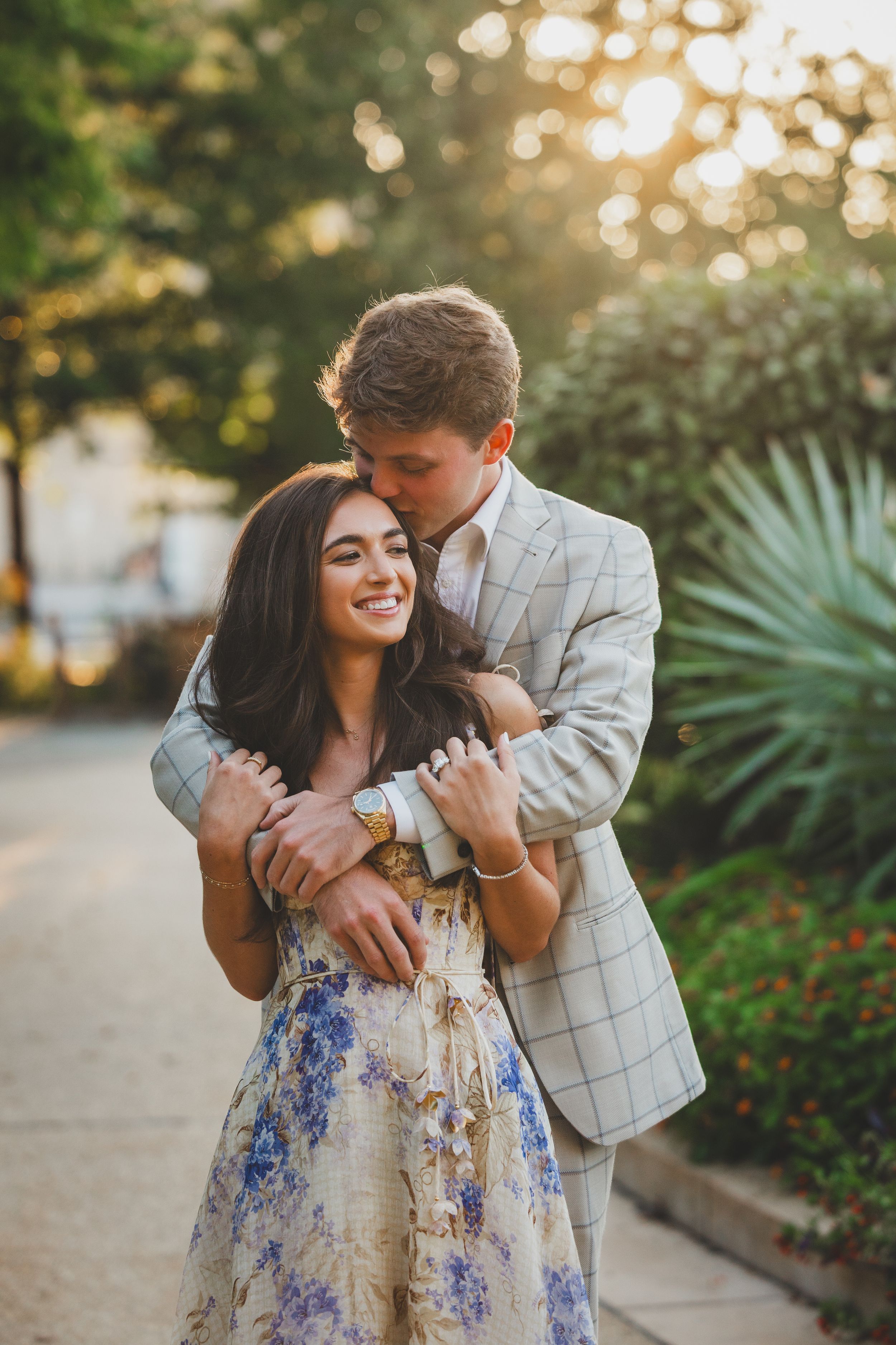 Romantic couple embraces outdoors at sunset, with woman in floral dress and man in plaid blazer.