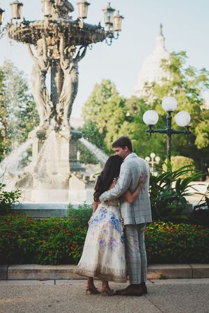 A romantic couple shares tender moments in a garden setting with ornate lamp posts and greenery in the background.
