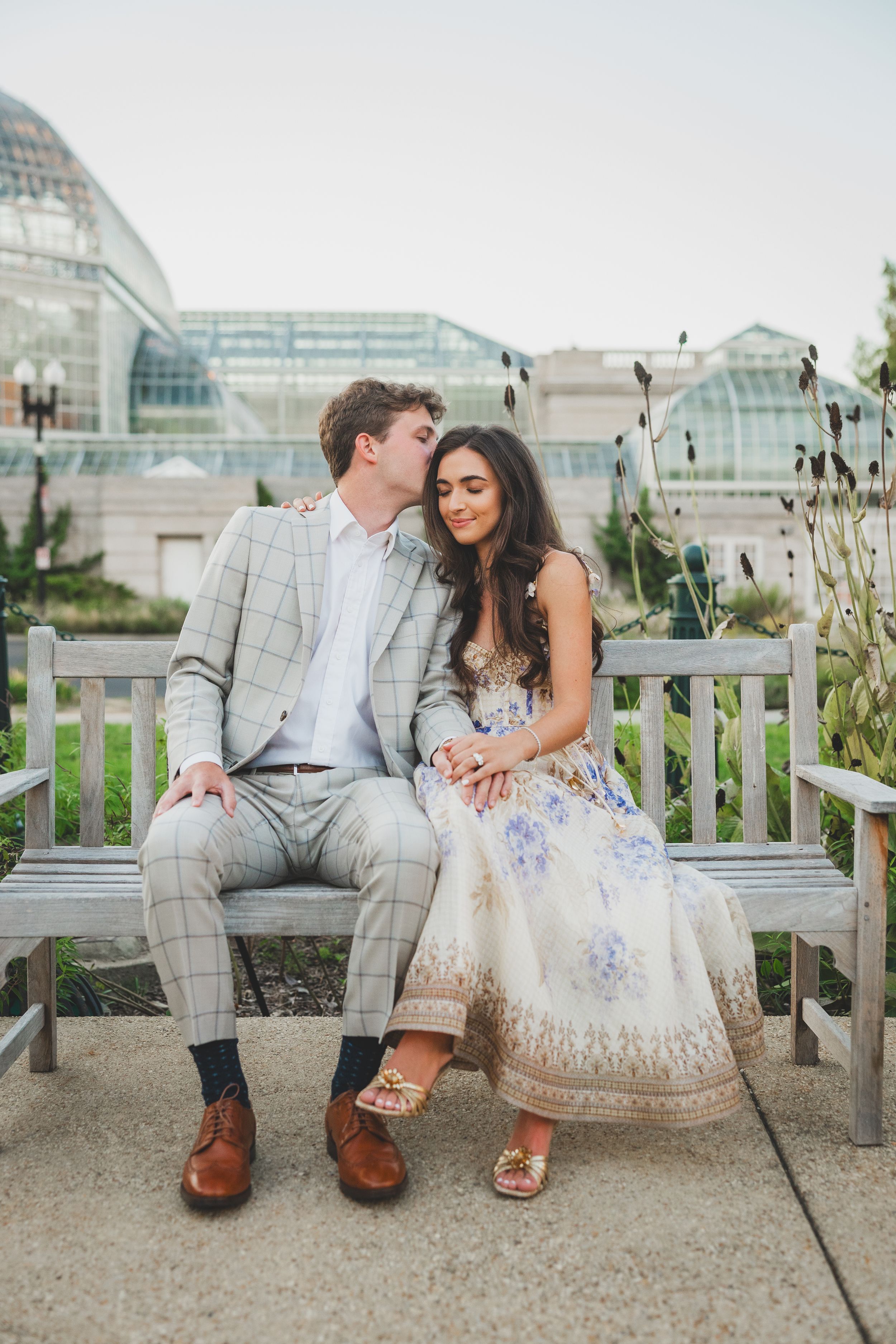 A couple sits together on a white bench in a romantic outdoor setting wearing coordinated formal attire at the Capitol Hill Gardens
