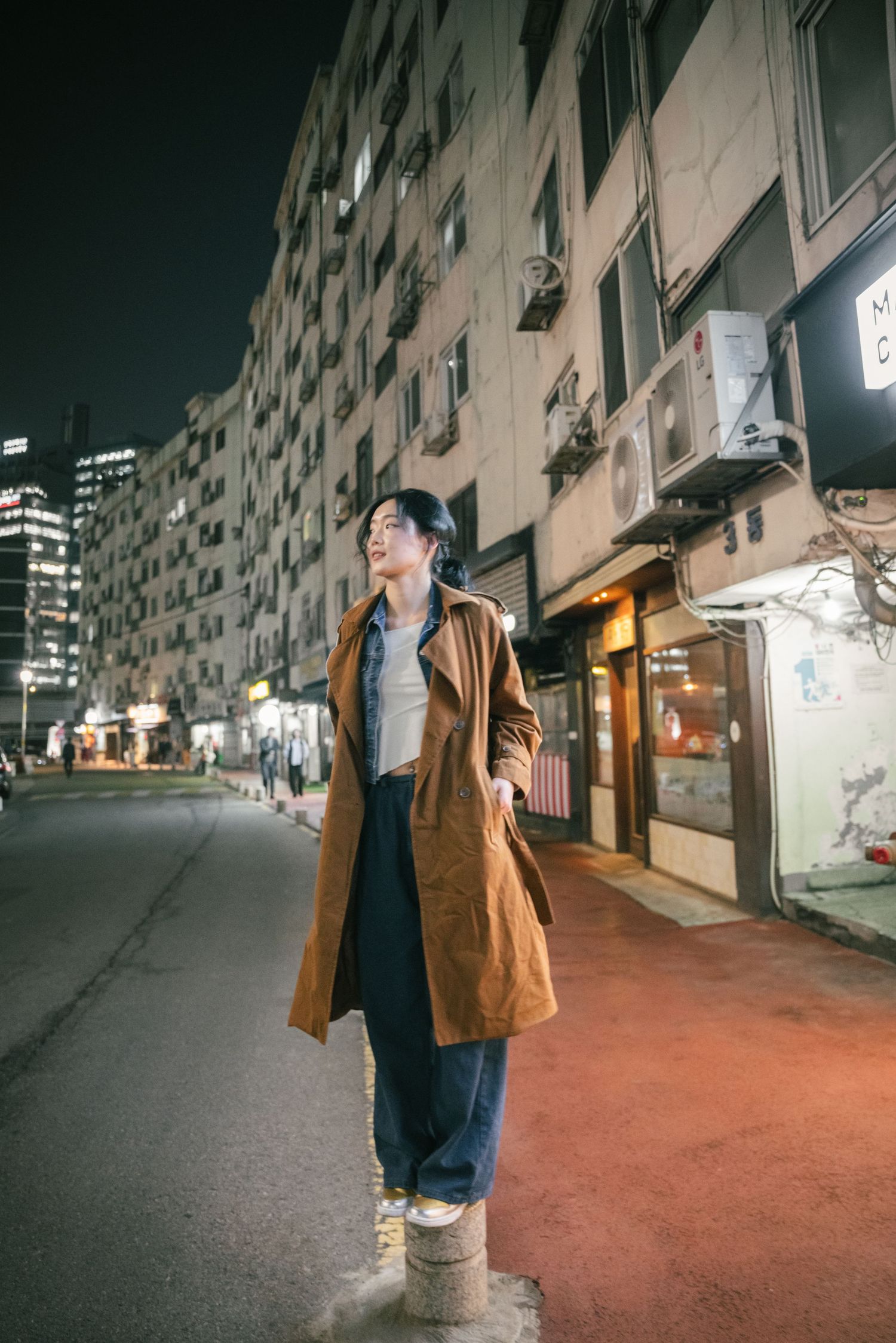 Person in brown coat stands on urban street at night with apartment buildings and air conditioners in background.