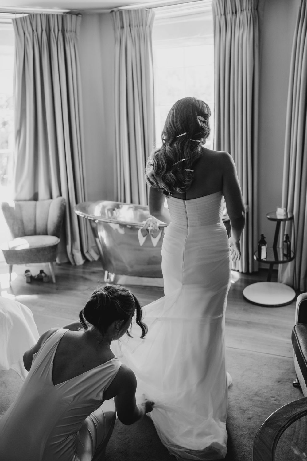 Black and white photo of final wedding dress preparations as someone helps adjust a white fitted bridal gown.