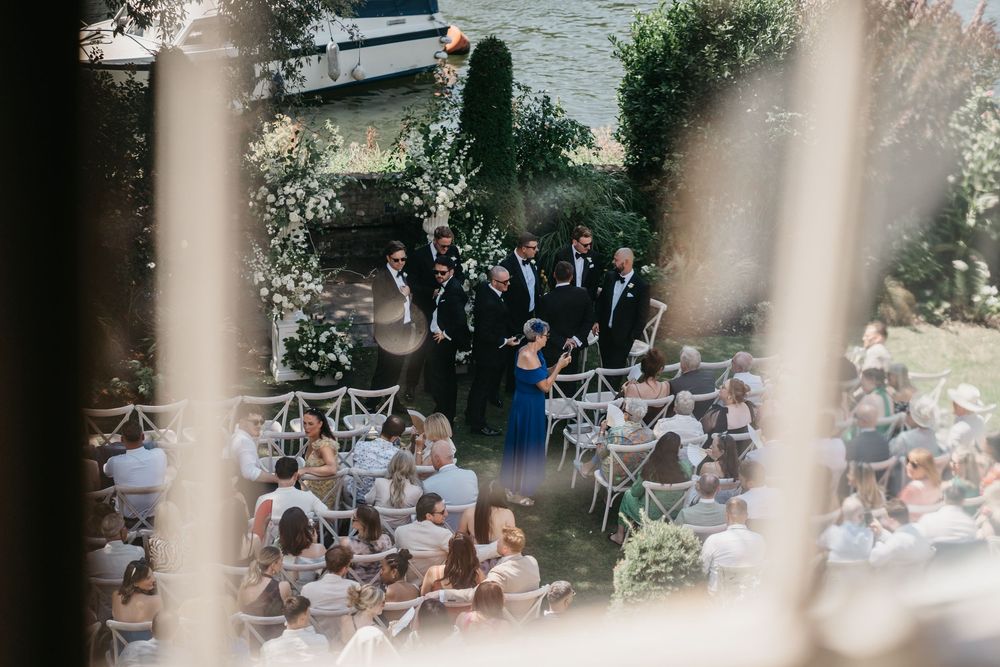 Outdoor wedding ceremony setup with white chairs arranged in rows under trees beside a waterfront venue.