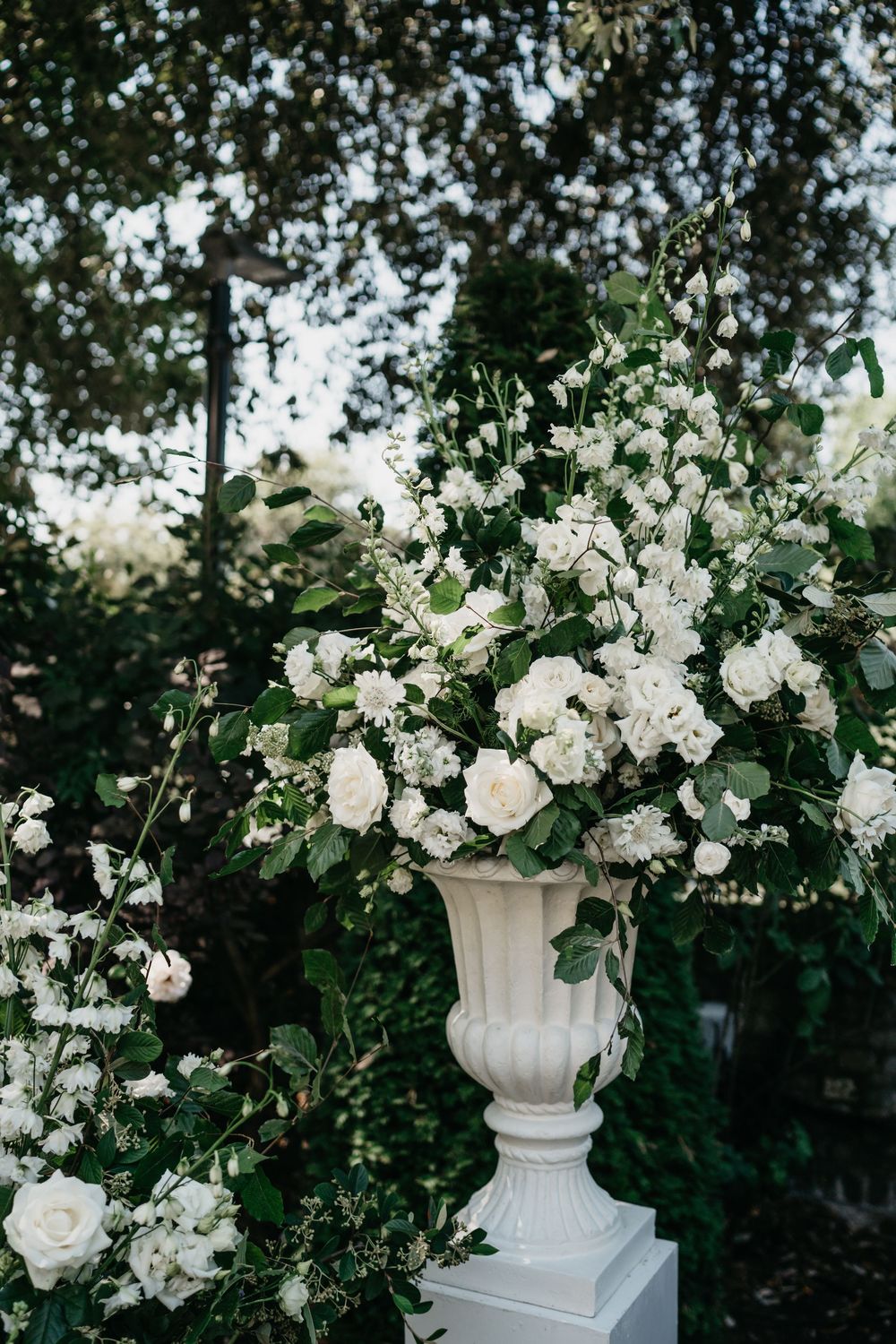 White roses and floral arrangements in elegant stone urns create a romantic garden setting against lush greenery.