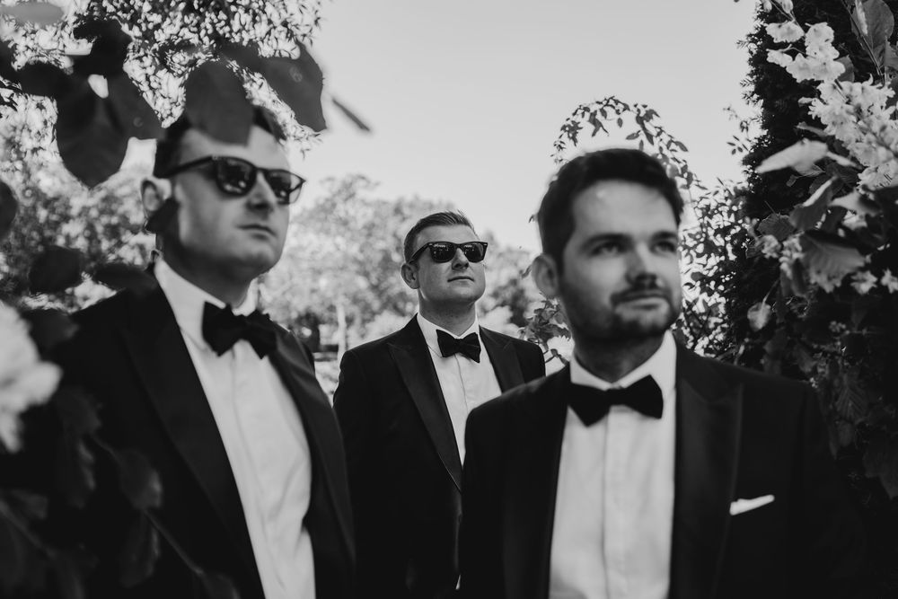 A group of formally dressed guests in sunglasses and tuxedos gather outdoors at an elegant event.