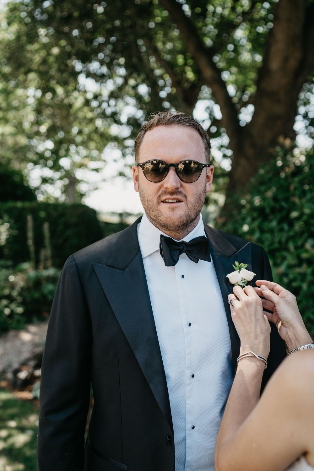A groom in a black tuxedo and bowtie gets ready for his wedding ceremony in an outdoor garden setting.