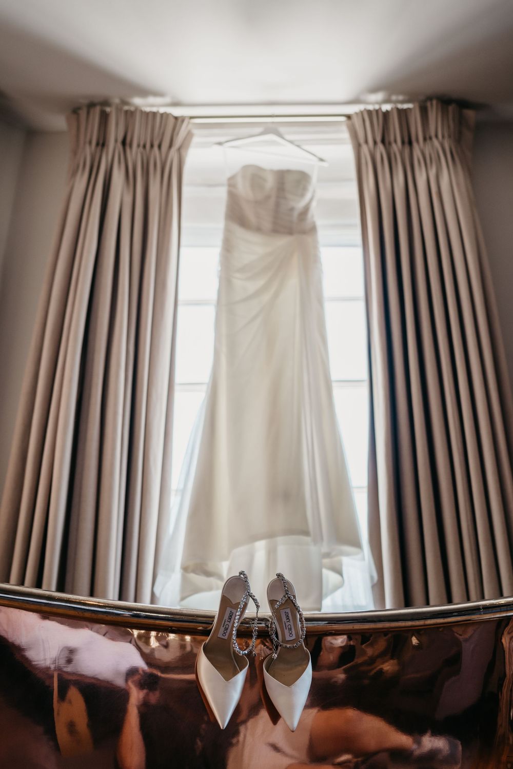 A wedding dress hangs in front of a window with elegant curtains while bridal shoes rest on the floor below.