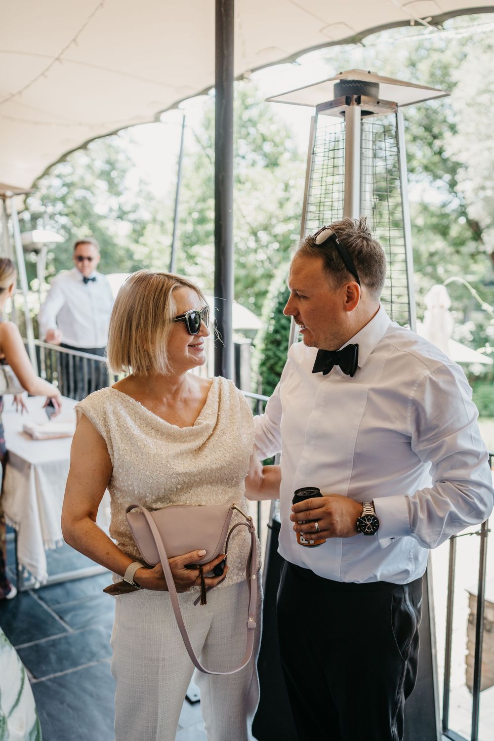 Two guests enjoying drinks and conversation at an outdoor wedding reception under a covered patio with a heat lamp.