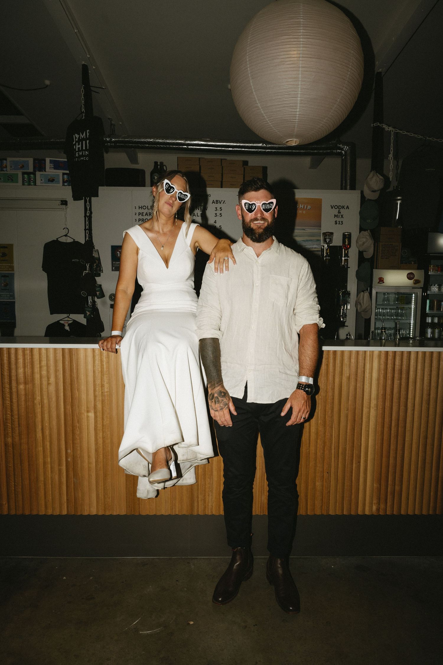 A couple poses at an indoor venue, with a large paper lantern above, the woman in white dress and man in casual attire.