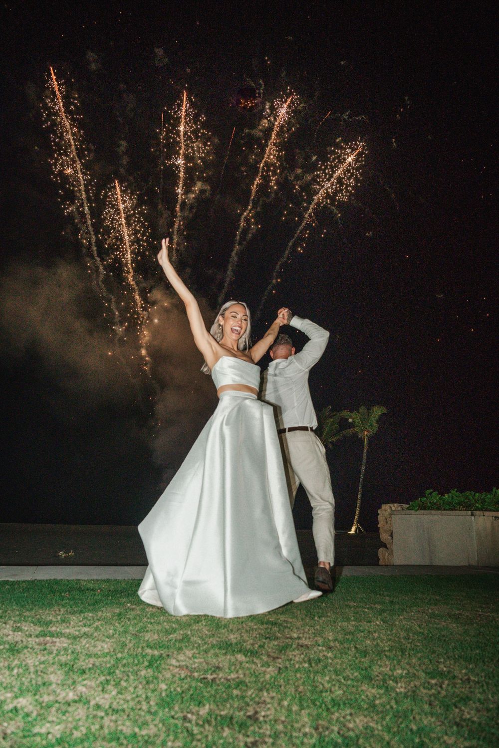 A couple in wedding attire celebrates with fireworks lighting up the night sky behind them on a grassy lawn.