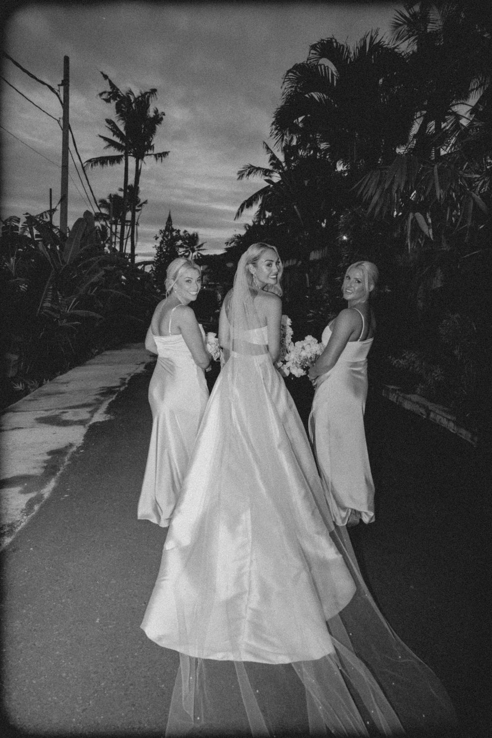 Three figures in flowing white dresses walk along a palm-lined path at dusk in a black and white photograph.