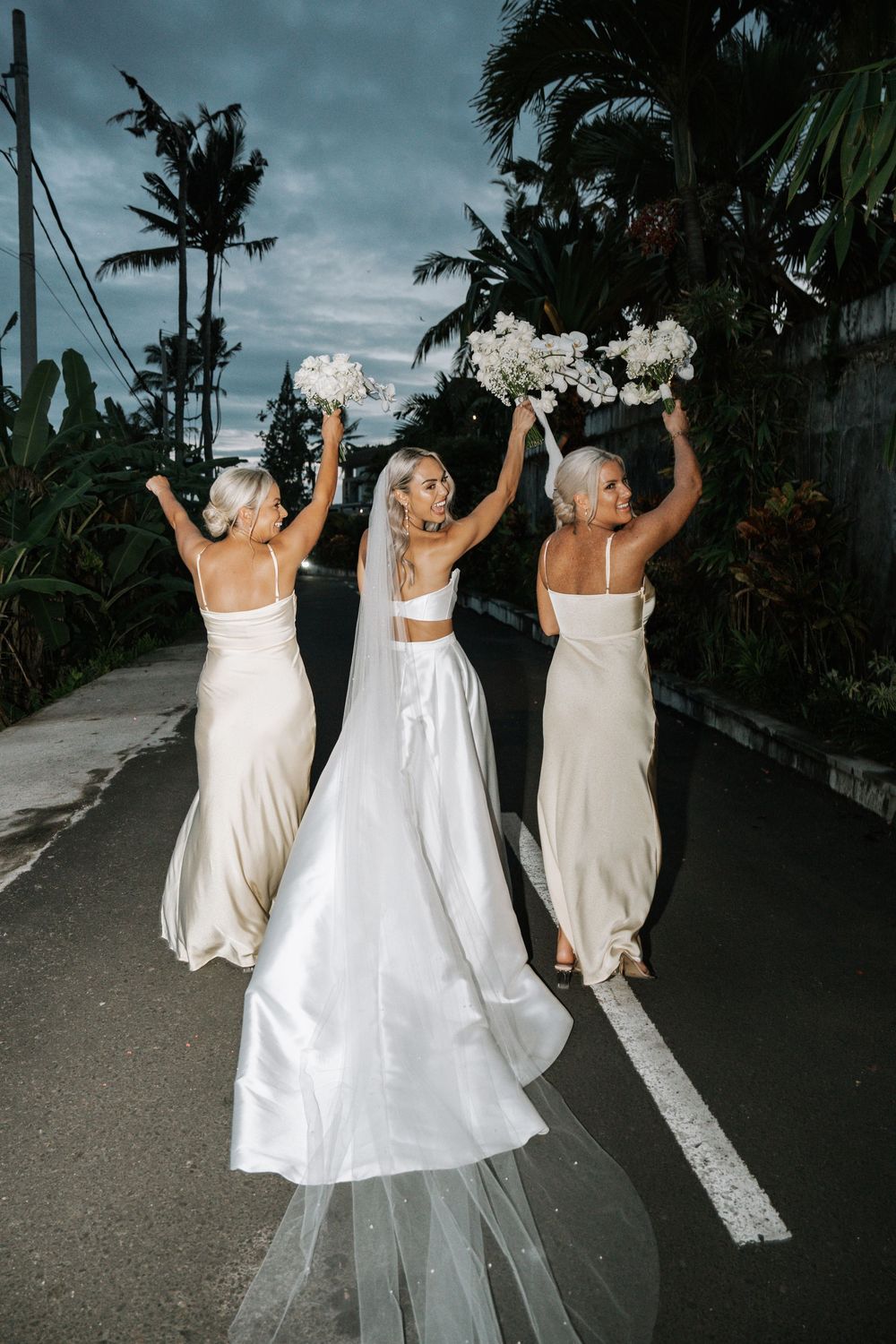 Three people in white dresses jump joyfully on a dark road with palm trees in the background.