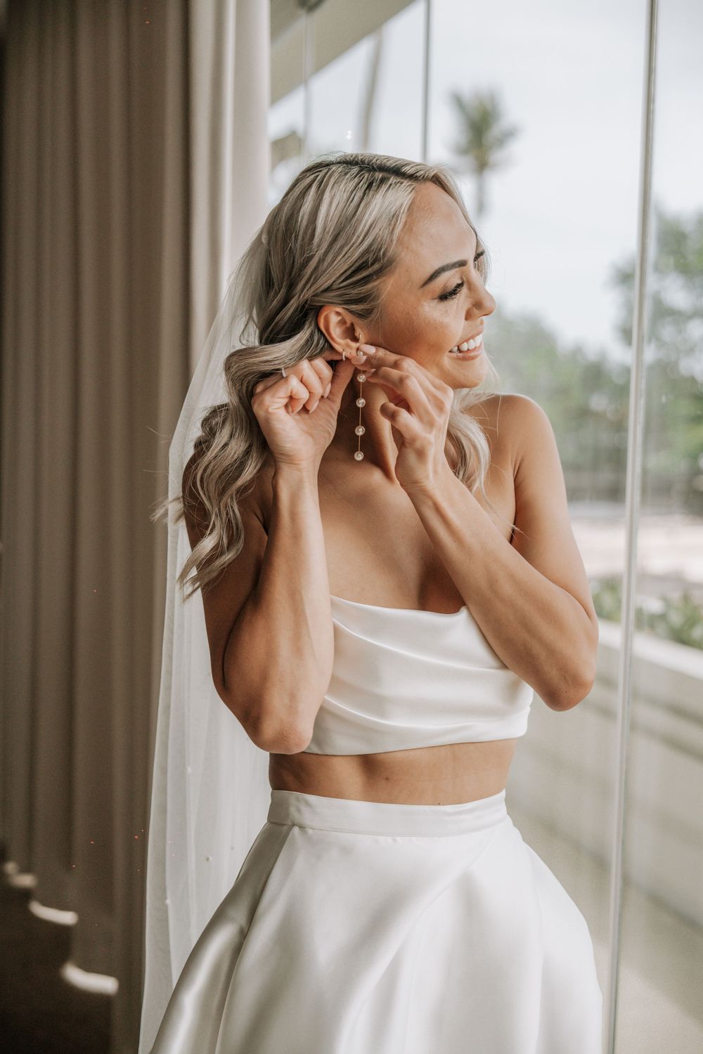 A bride in a white wedding dress getting ready near a window with natural light streaming in.