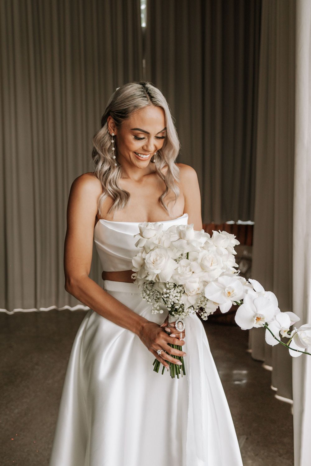 A bride in an elegant white wedding dress smiles while holding a beautiful bouquet of white orchids and baby's breath.