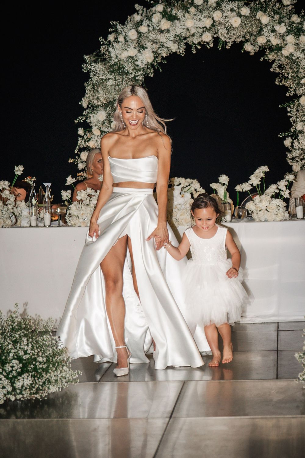 Two figures in white dresses walk down steps under a floral arch at an elegant wedding reception.