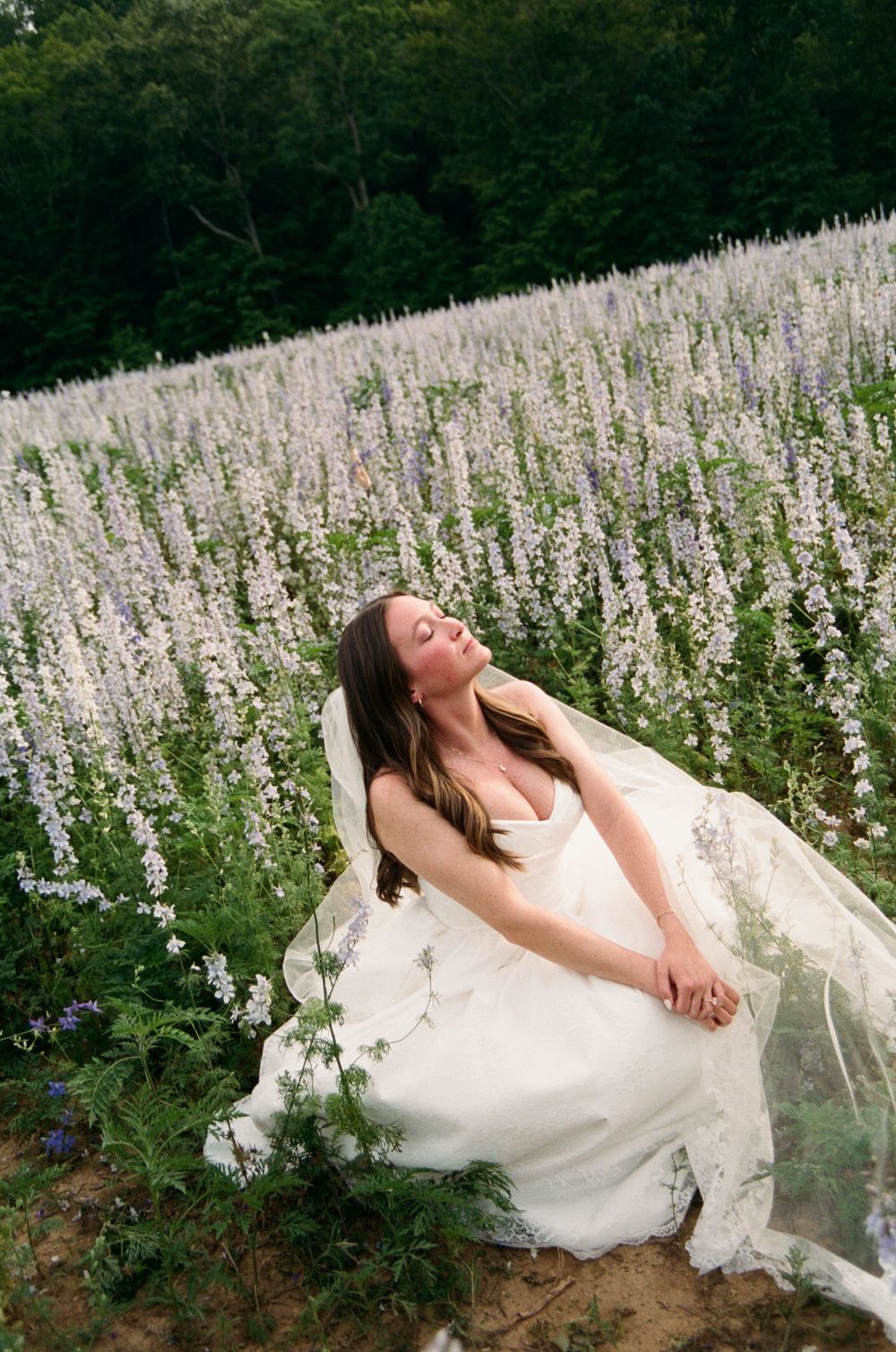 A bride in a white wedding dress poses gracefully in a field of delicate white wildflowers on a summer day.