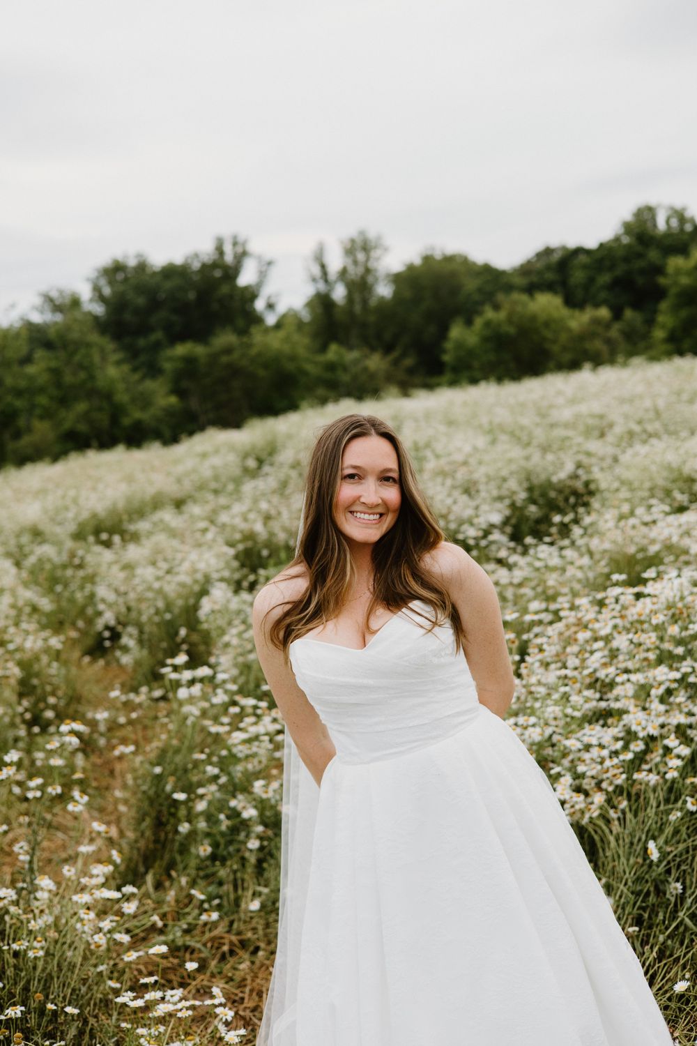 A bride poses playfully in a white gown among a meadow of wild daisies during a summer photoshoot.