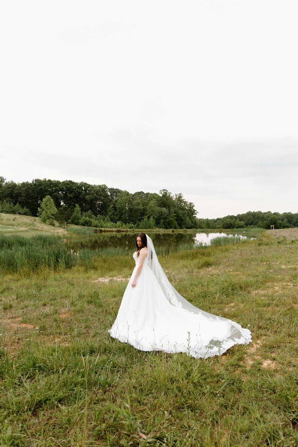 A flowing white wedding dress with a long train spreads across a grassy hillside on a cloudy summer day.