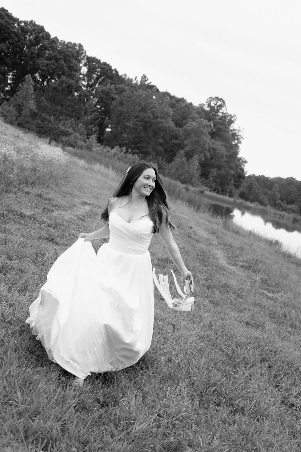 A person in a flowing white wedding dress twirls in a grassy field near a lake on a summer day in black and white.