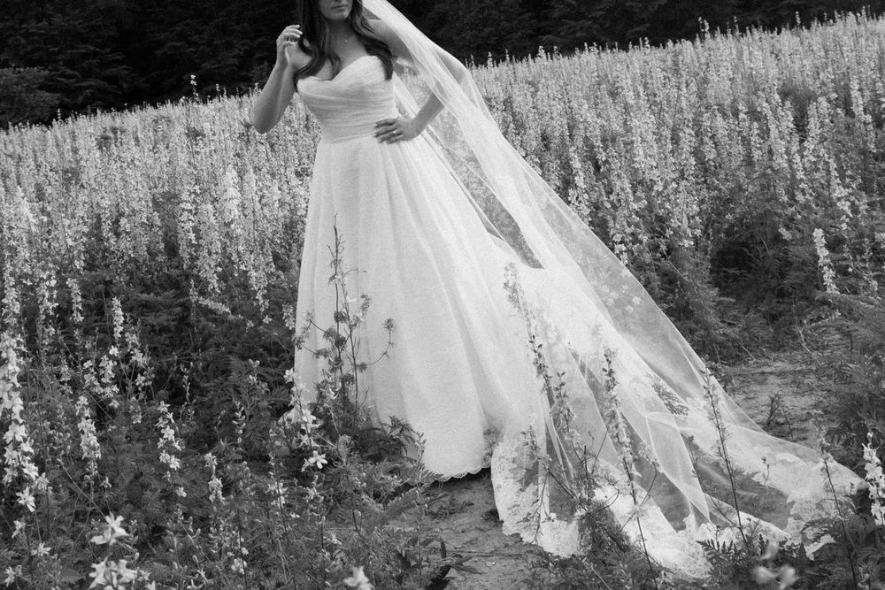 A flowing white wedding gown with long train trails through a meadow of wildflowers in black and white photography.