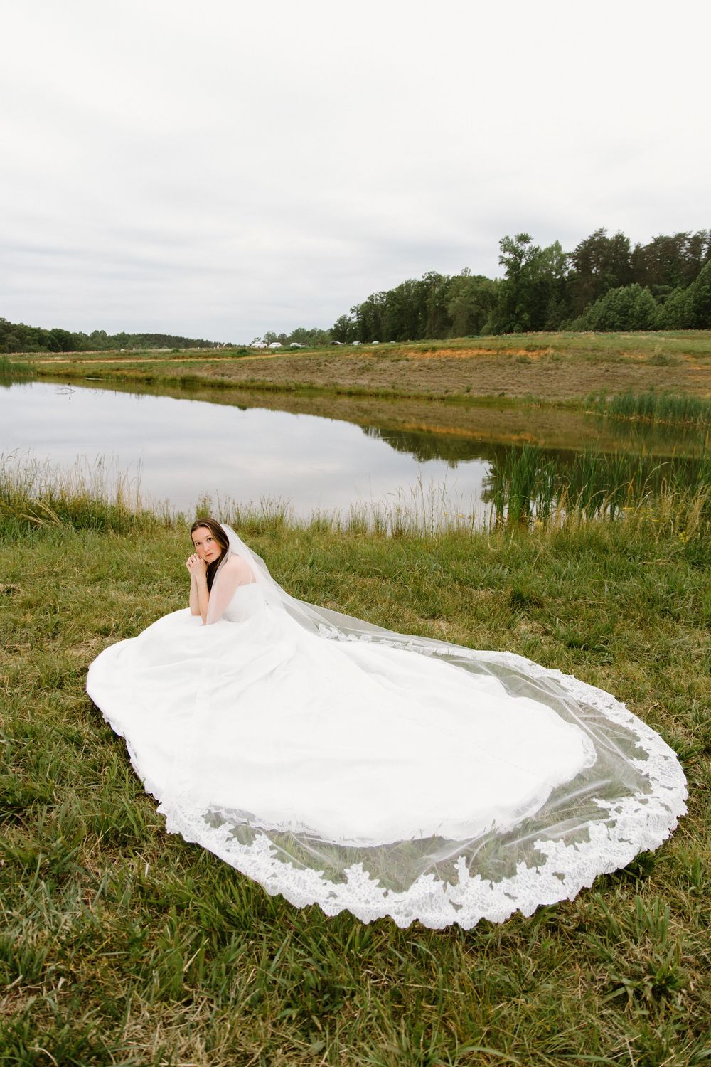 A flowing white wedding dress with lace details spreads across a grassy field with a lake and trees in the background.