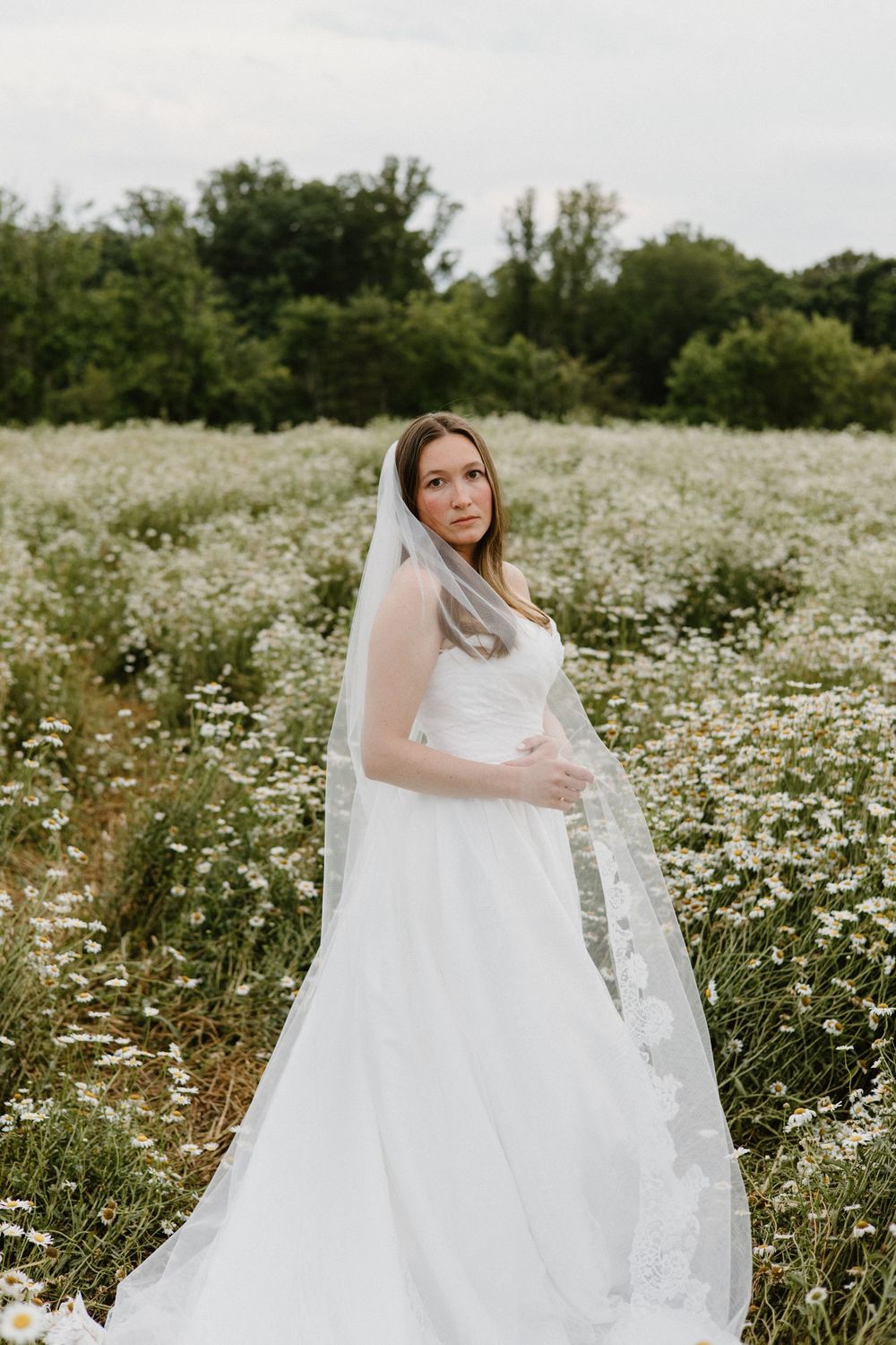 A bride in a flowing white wedding dress stands in a field of blooming white daisies on an overcast day.