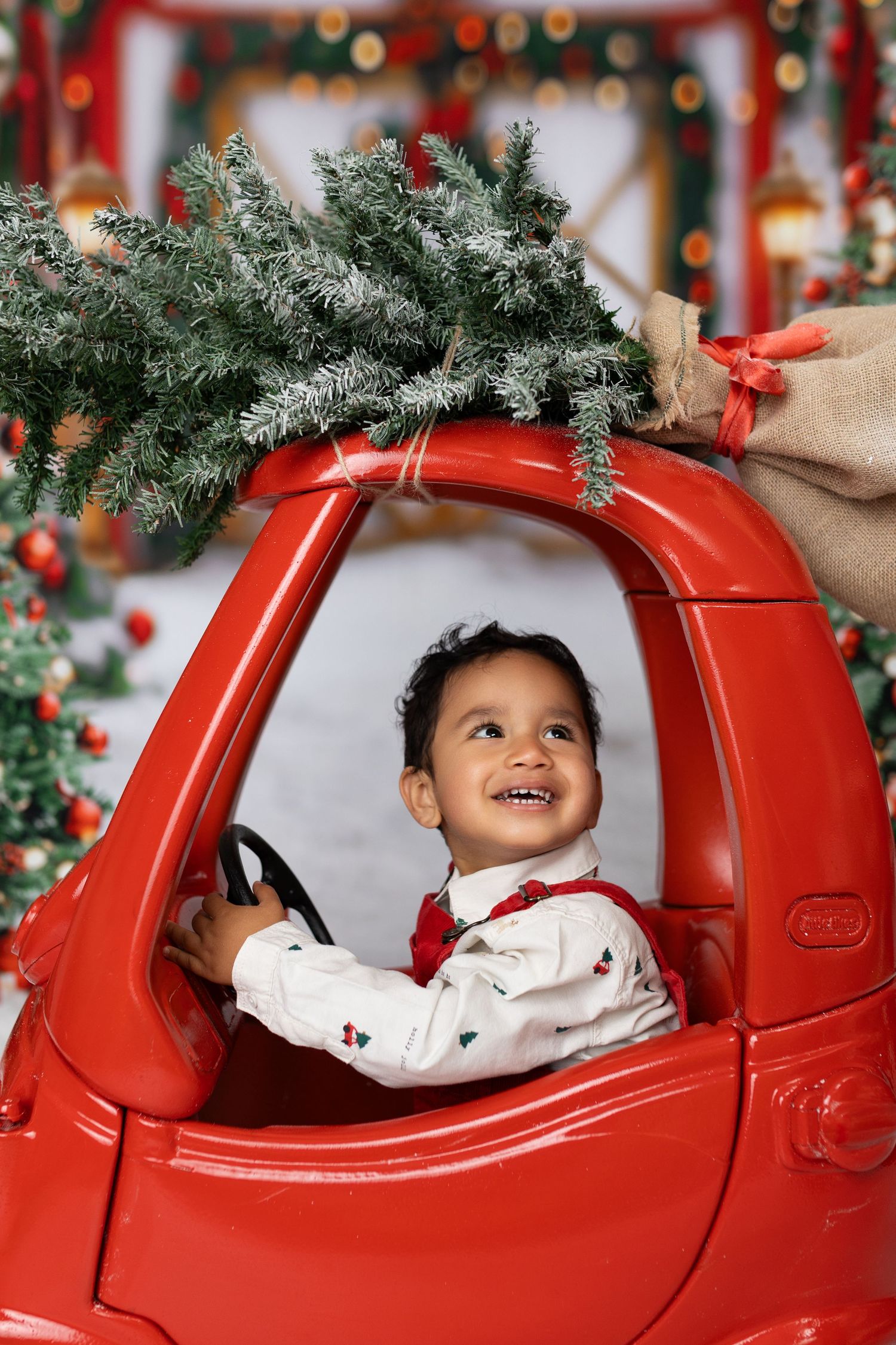 A child sits in a bright red sleigh against a snowy backdrop with Christmas trees and twinkling lights.
