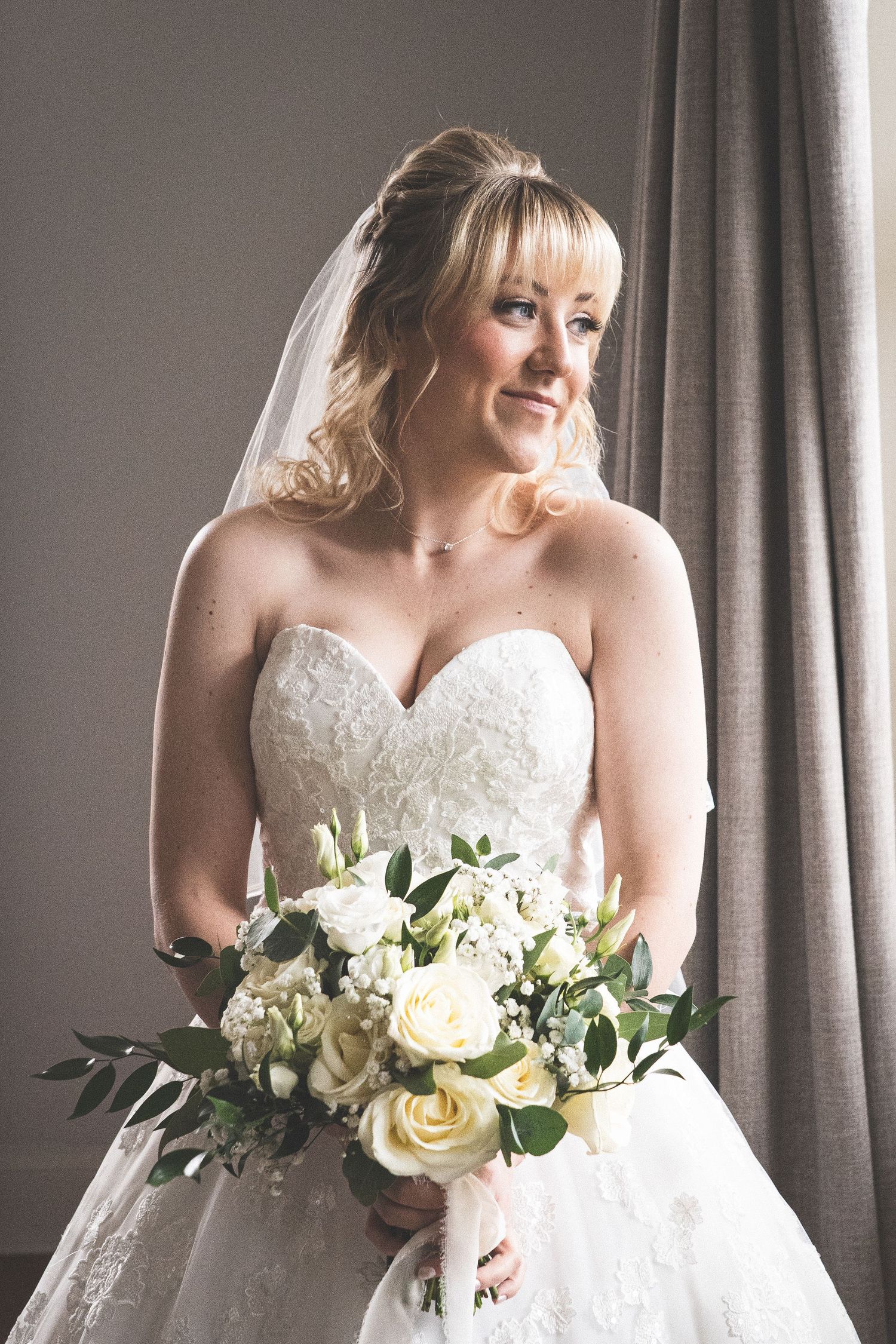 A bride in an elegant lace ball gown holding a white floral bouquet in a sunlit room.