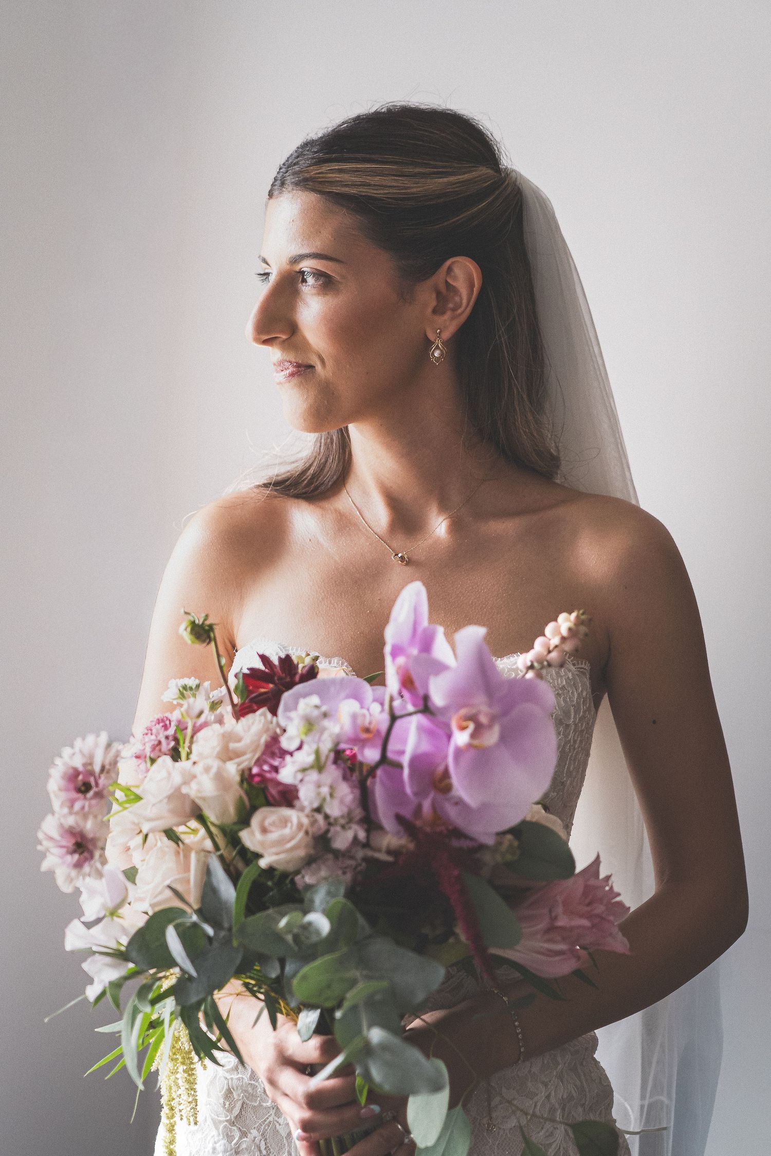 A bride in an elegant lace ball gown holding a white floral bouquet in a sunlit room.