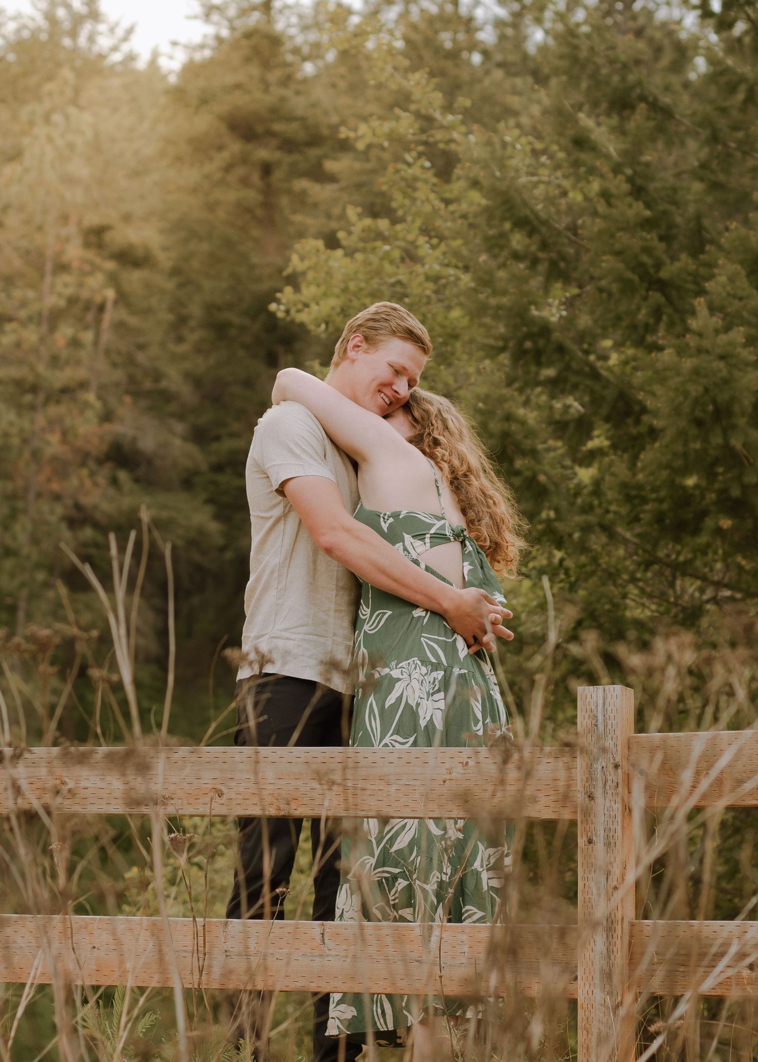 Couple embracing happily in Northern Idaho among Pine trees. Man wearing khaki shirt and black pants. Woman wearing a green leaf lined dress