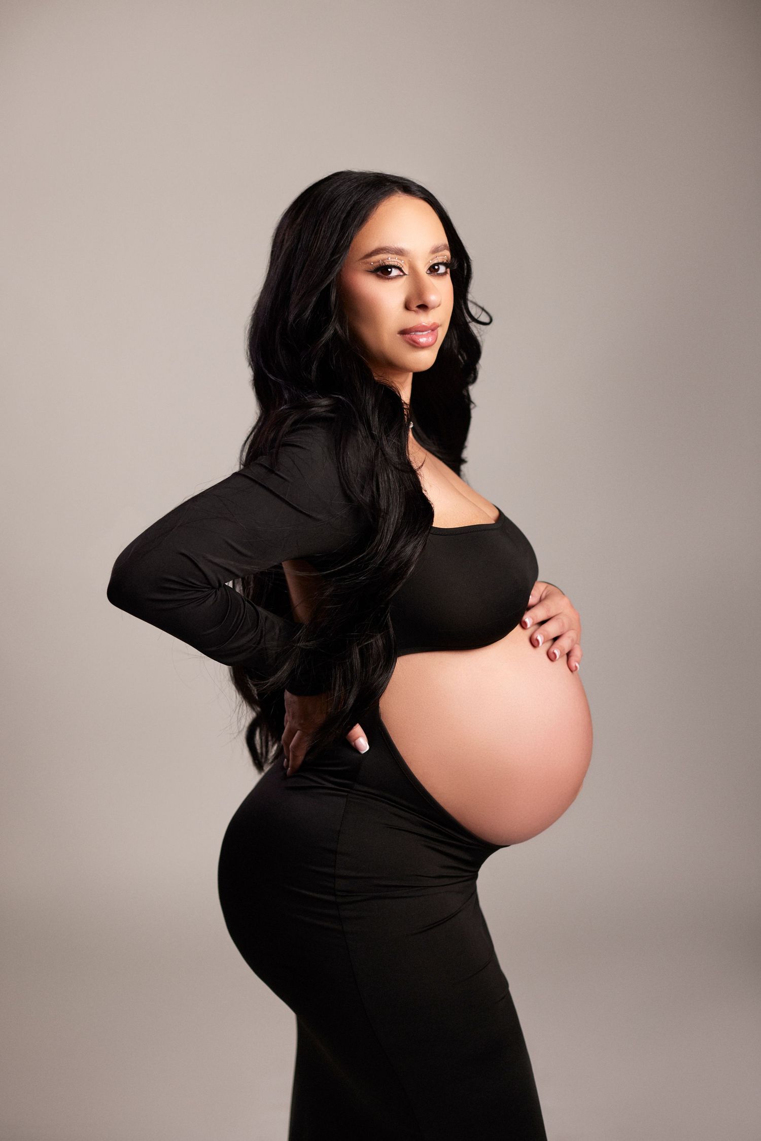 Maternity photoshoot featuring a pregnant woman in a black dress against neutral backdrop.