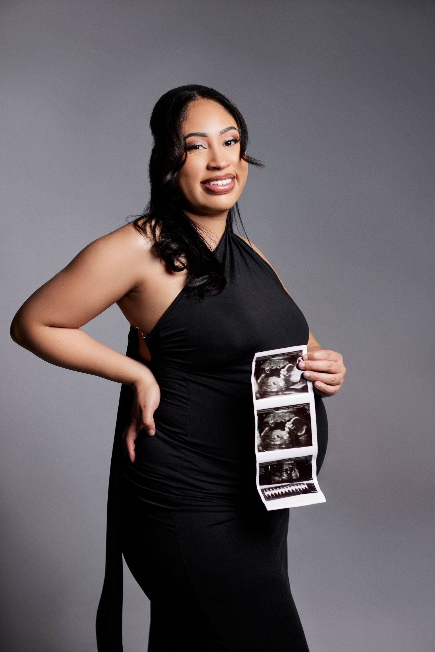 Expectant mother in black dress holds ultrasound photos during pregnancy photoshoot.