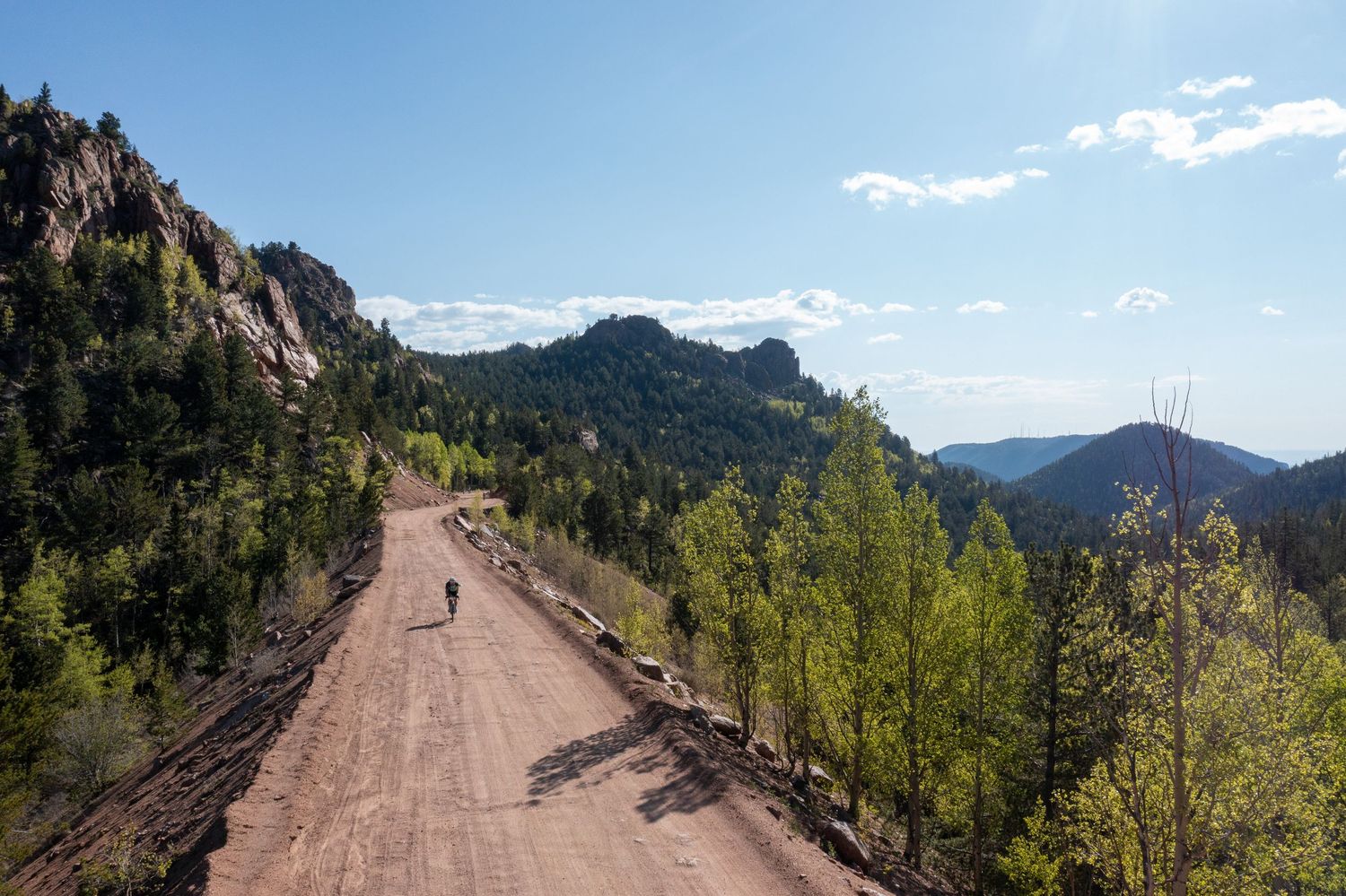 A motorcyclist rides on a dirt road through a forested mountain landscape with bright sunlight overhead.