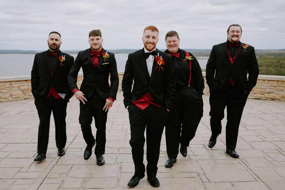 Groomsmen posing playfully in black suits against an overcast sky.