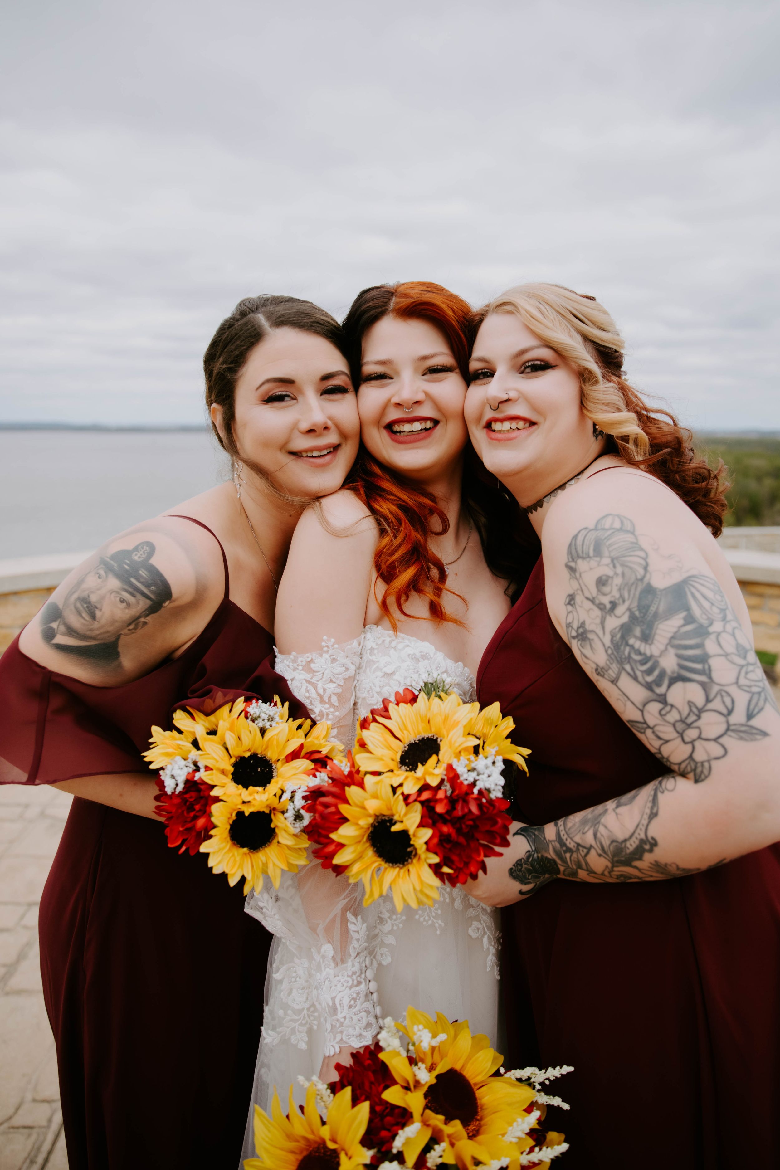 Bridesmaids in burgundy dresses holding sunflower bouquets pose together outdoors.